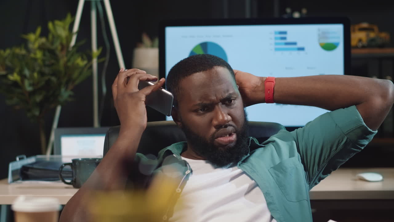 Focused african american hipster guy talking cellphone in night office.