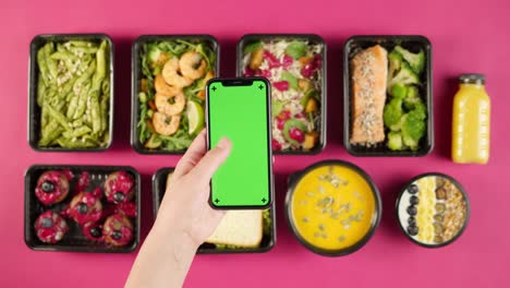 food delivery top view, take away meals in disposable containers on pink background. lunch boxes with cooked vegetarian dishes, using phone with chroma green screen. healthy diet. catering service