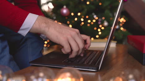 close up view of man hands typing on laptop sitting near the christmas tree in living room with christmas decoration