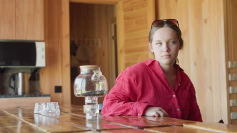 woman in red shirt seated at wooden table in warm cabin kitchen gazes thoughtfully at transparent glass kettle stacked on glasses, morning light fills rustic space with wooden walls