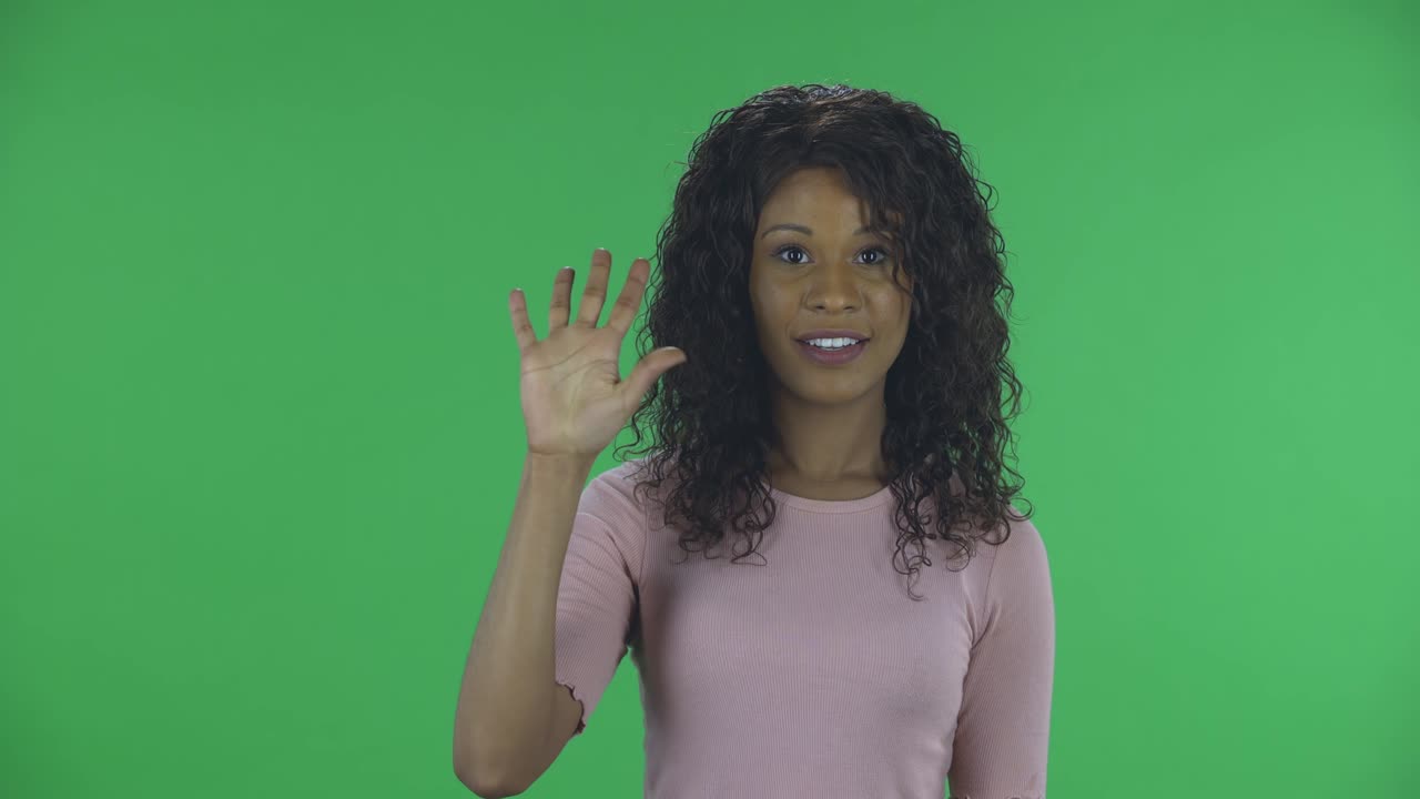 Portrait of beautiful african american young woman is looking at camera and shows fist, then counting one, two, three, four, five fingers. Burning brunette with wavy hair in jeans and a beige blouse on a green screen in the studio