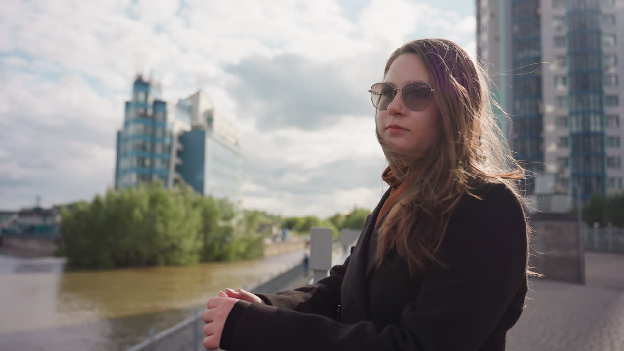Lady stands opposite river wearing winter coat and eyeshade gazing in distant direction with calm reflection, soft light illuminating her face against modern building and blue sky creating peace