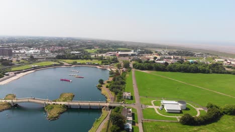 Drone-Elevándose-Sobre-El-Lago-Sobre-La-Ciudad-Costera,-Gente-Cruzando-El-Puente-En-Un-Día-Soleado