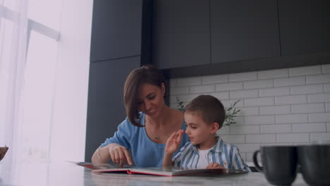 una hermosa madre y su hijo sentados en una mesa en una cocina luminosa leyendo un libro y mirando imágenes metiendo un dedo en un libro y hojeando las páginas