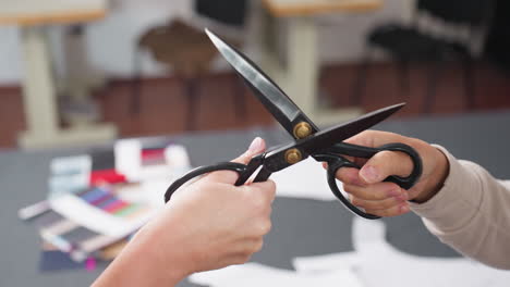 female hands holding black and golden color scissors, engaging in playful sword fight above work table filled with sewing patterns and colorful fabric samples in creative bright workspace