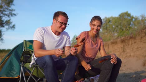 Couple enjoying grilled sausages at a campsite by a river