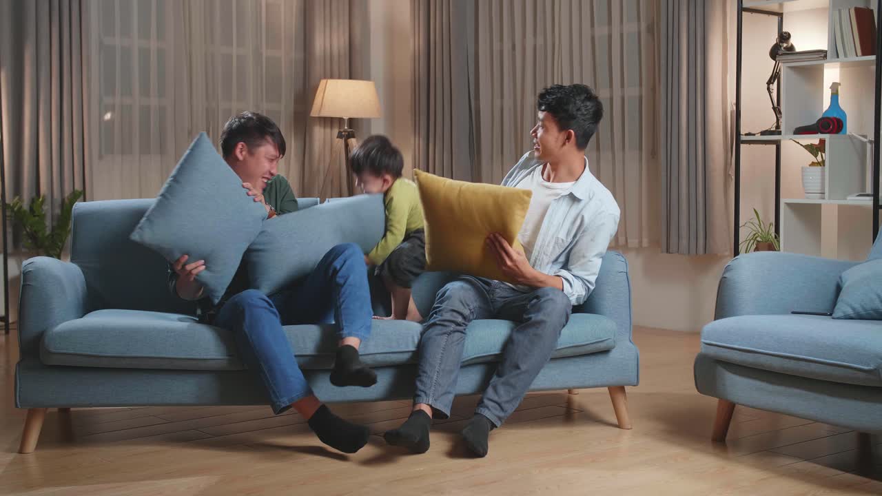 Happy Asian Gay Couple Having Fun With Their Lovely Son Fighting Pillows In Living Room
