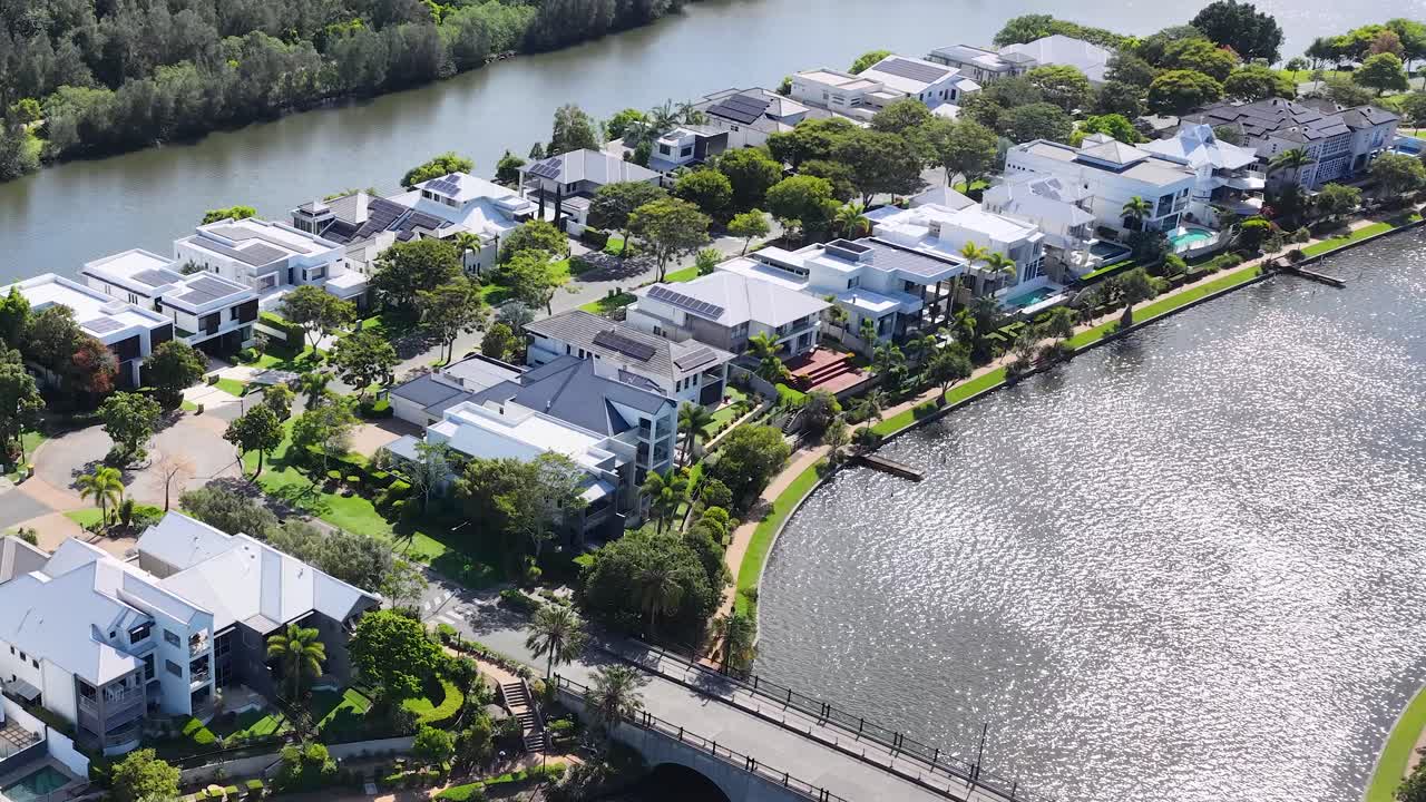 Drone glides above sunny canal neighborhood, revealing modern waterfront houses, bridge, and lush greenery