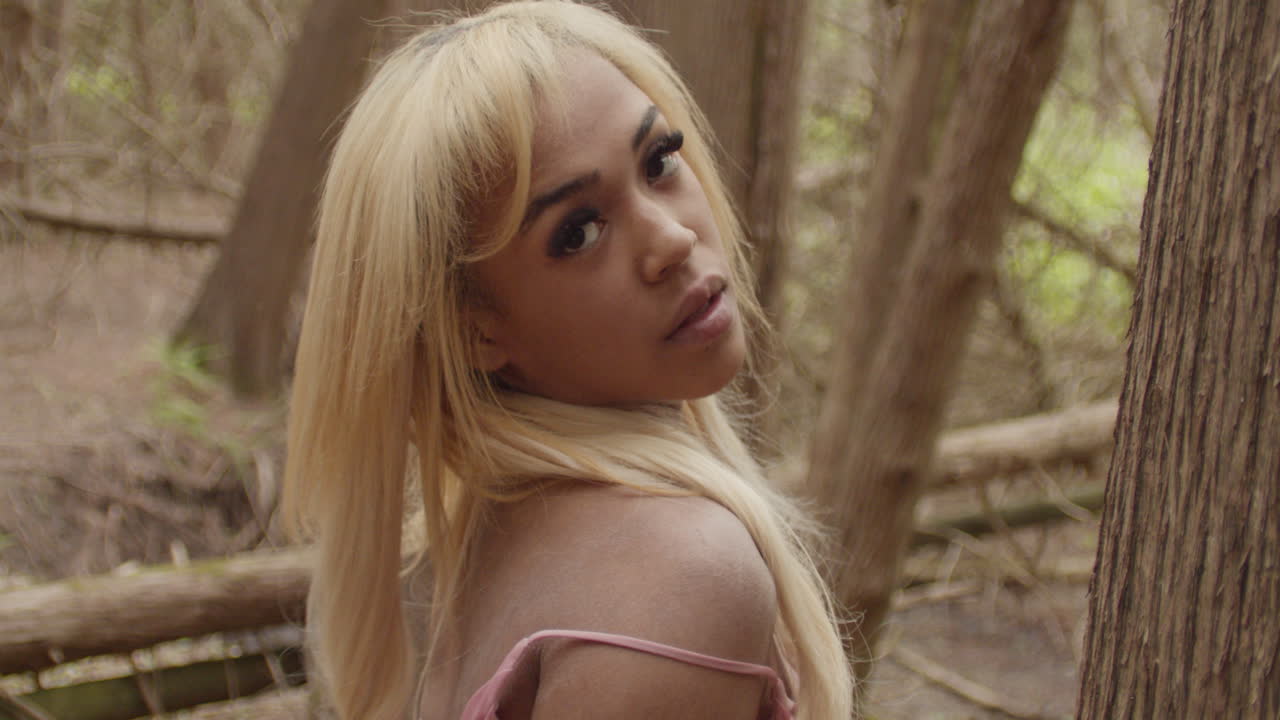 Young ebony woman in pink gown. Handheld camera in close up tilts from her hands to her face . She flips her hair before looking seductively at camera. She looks upward to the sky. 60fps slow motion.