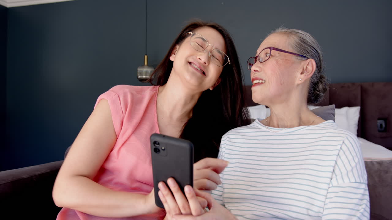 Holding smartphone, asian grandmother and granddaughter laughing together on couch