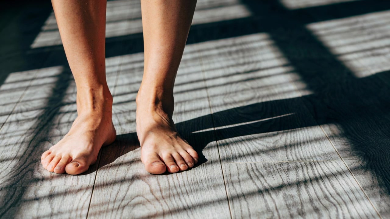 Shifting bare feet and lower legs making subtle weight shifts on wood floor, blinds casting stripes