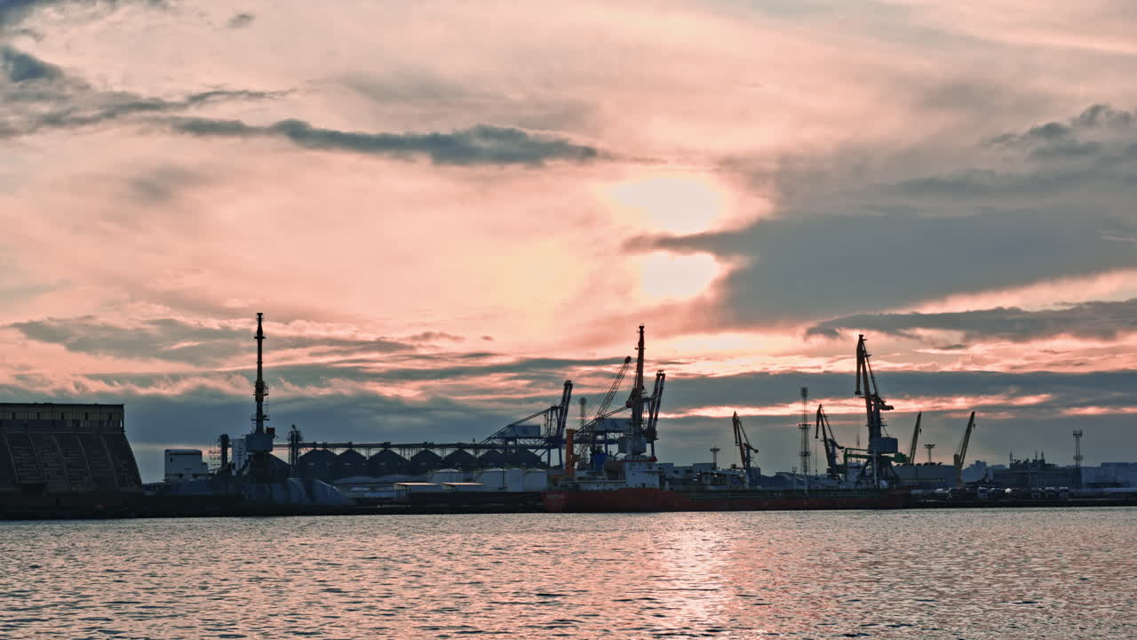 Sunset view of a busy port with cranes and ships at work, showcasing the hustle of evening operations near the water, with clouds reflecting in the sky