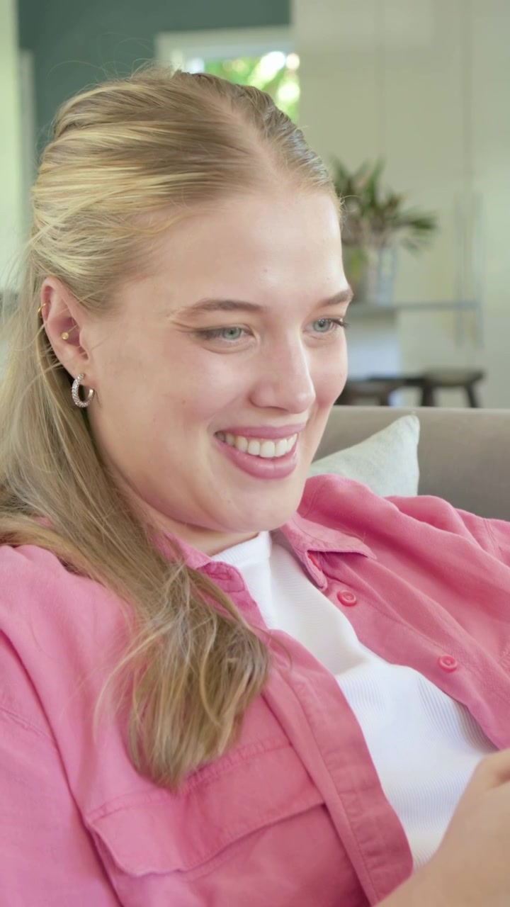 Vertical video: Cradling pink mug, woman reaching blue phone, reading, smiling on couch in pink top
