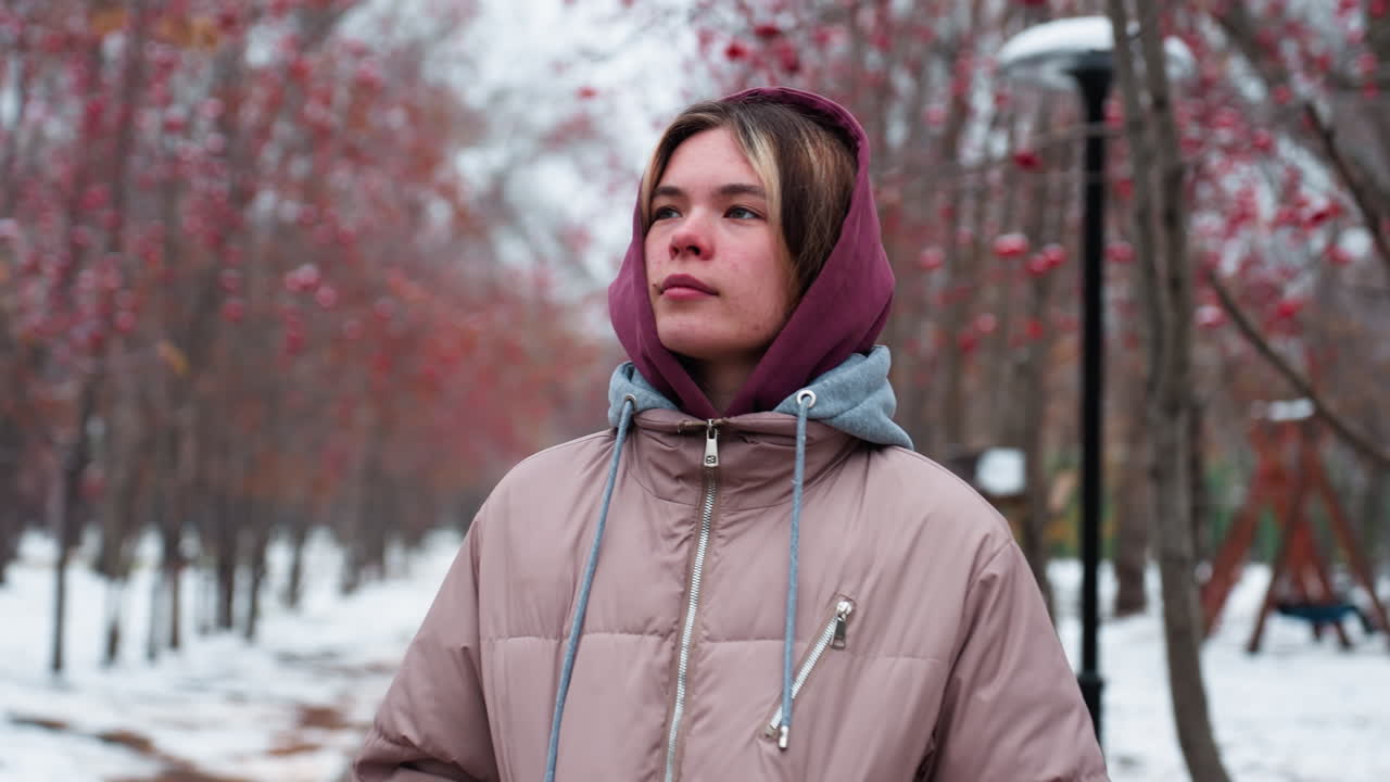 따뜻한 후드 재을 입은 젊은 여성은 은 베리 나무로 둘러싸인 눈으로 인 길을 따라 고, 주위를 상하며, 배경에는 멀리 흐릿한 인물이 있습니다.