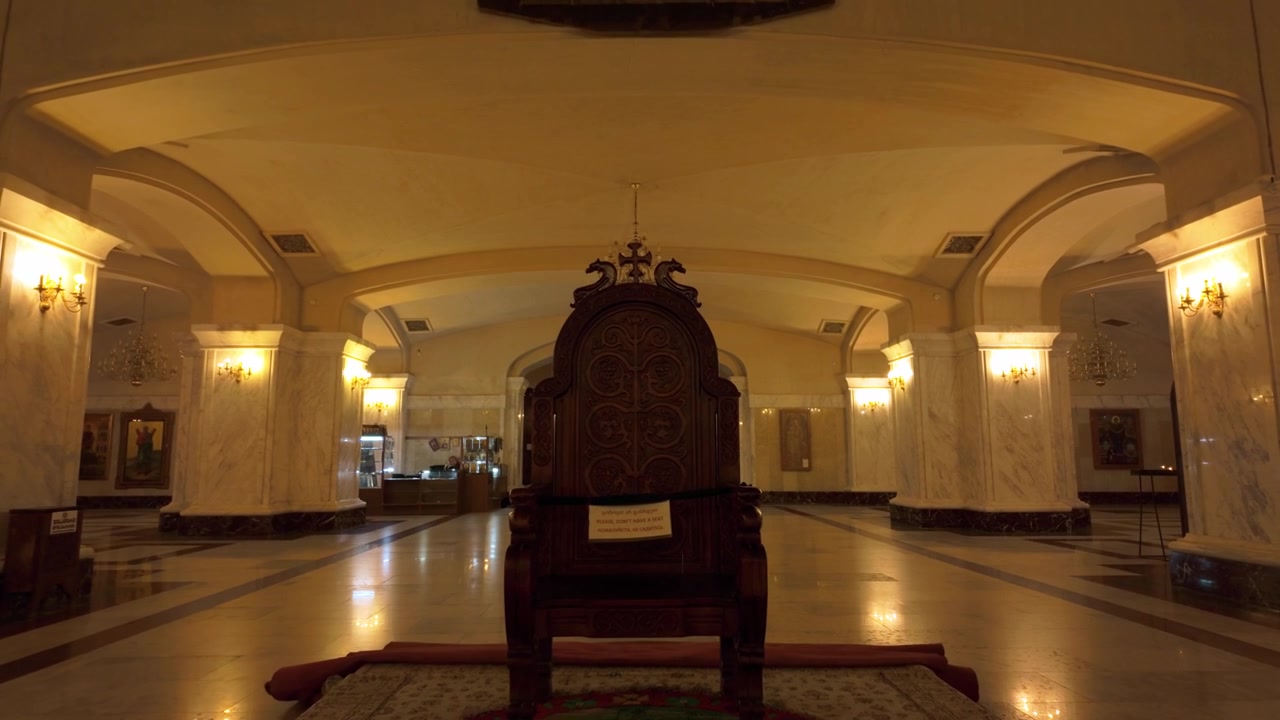 Trono in legno intarsiato posizionato centralmente nella vasta sala della Cattedrale della Santissima Trinità a Tbilisi. Pilastri in marmo, arcate e lampadari d'ambiente definiscono lo spazio sacro