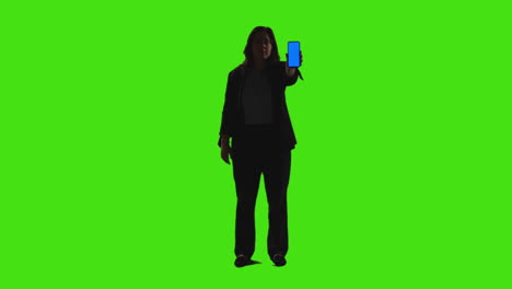 full length shot of businesswoman holding blue screen mobile phone towards camera standing against green screen with low key lighting