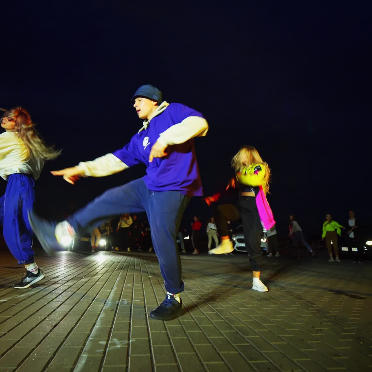 Teenage boy and girls dancing hip hop outdoors at night. Low angle view. People at backdrop dancing near the car with lights on