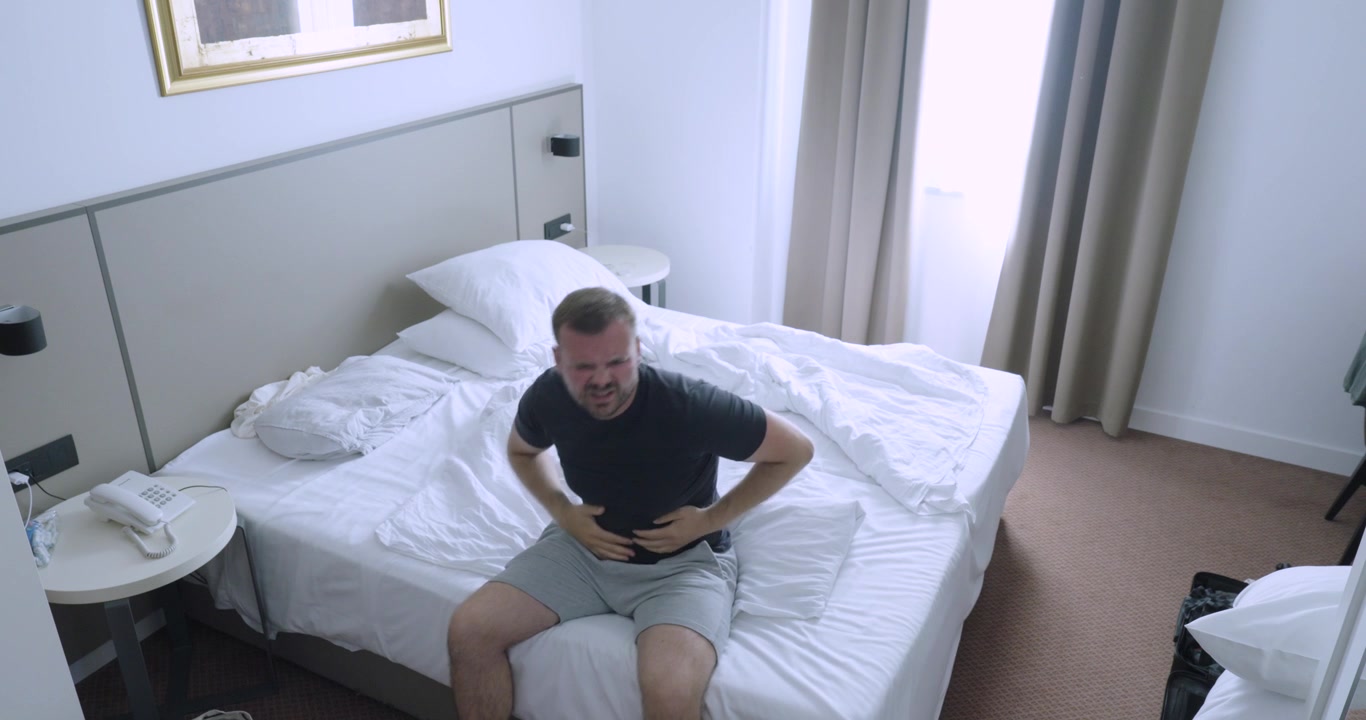 Uomo seduto su un letto d'hotel che si sente male durante la sua vacanza. Concetto di viaggio che include malattia, influenza, disagio, recupero e piani di vacanza interrotti