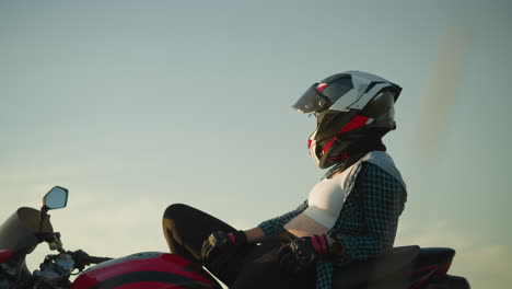 a female biker is seen resting on her red power bike, wearing a helmet and gloves, as she looks forward into the open field