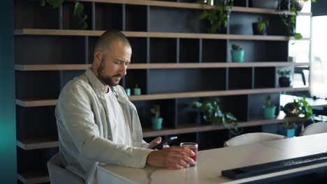 Happy-bearded-man-looking-at-mobile-phone's-screen-and-smiling,-cocktail-in-hand