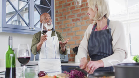 Una-Pareja-De-Ancianos-De-Raza-Mixta-Usando-Delantales-Hablando-Mientras-Cocinan-Juntos-En-La-Cocina-De-Casa