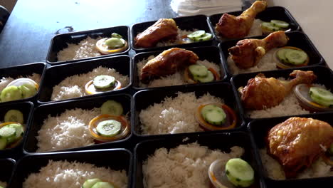 a row of nasi lemak in lunch boxes container while hands put a chicken roasted on it