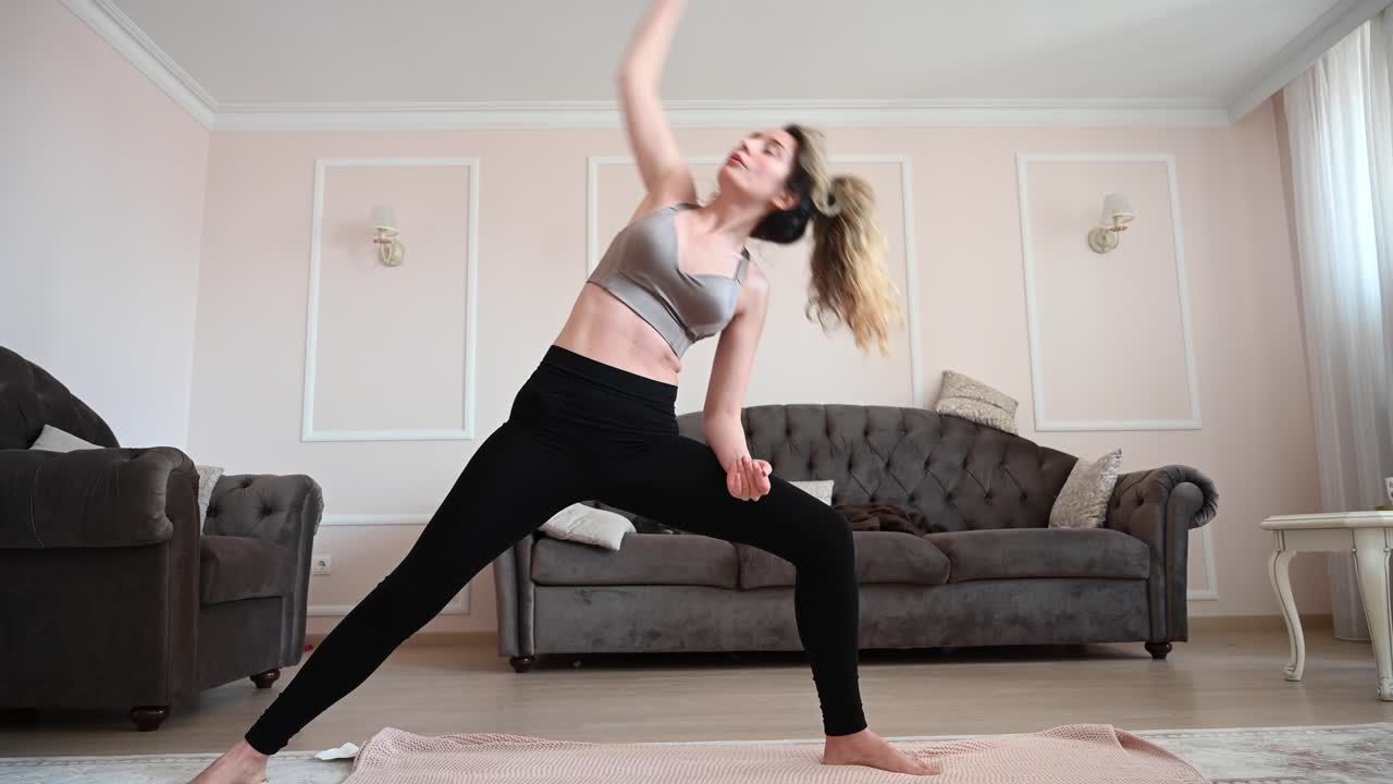 Young woman sitting cross legged on the floor in a bright living room, practicing yoga meditation with arms raised and eyes closed
