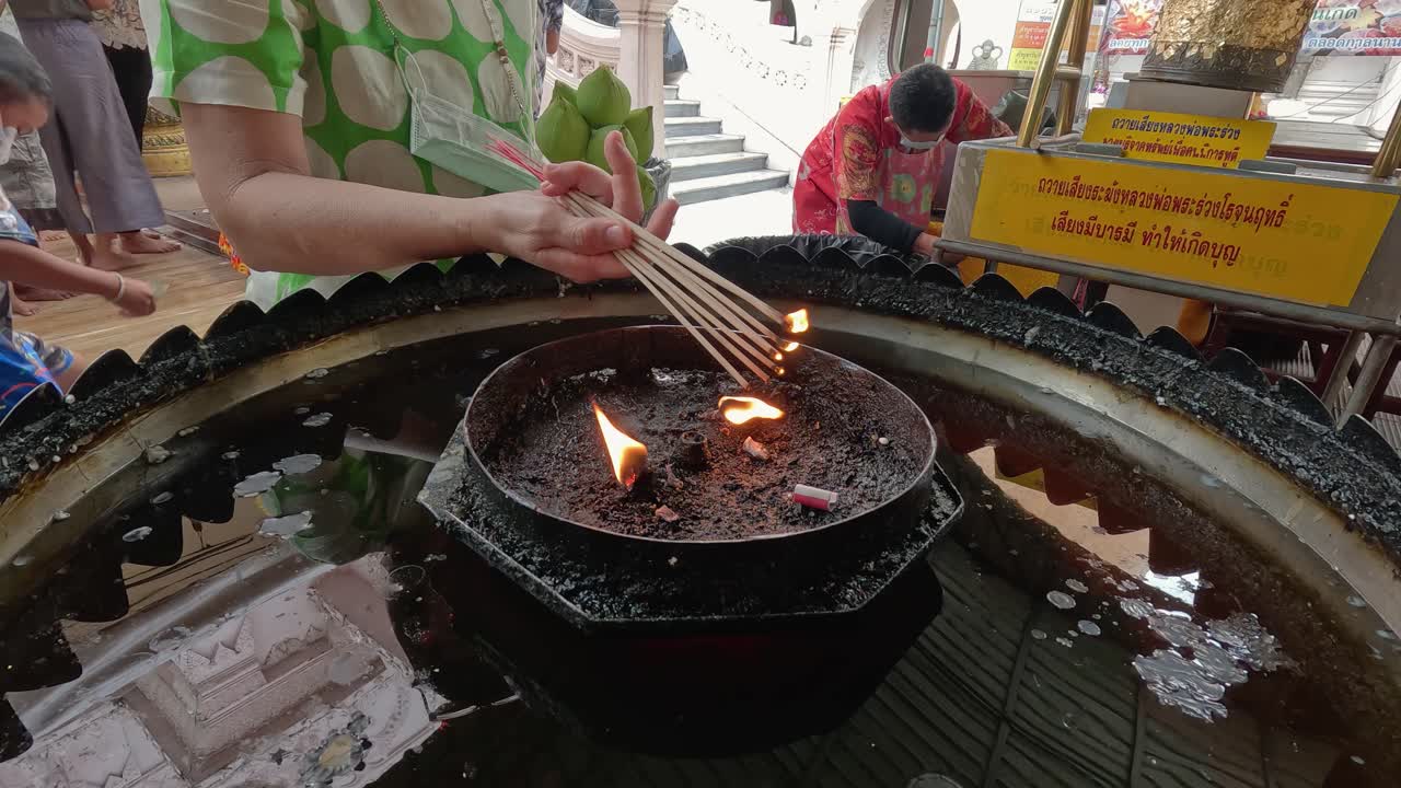 A person lights incense sticks over a flame in a temple setting, with natural daylight, close-up composition, and subtle camera movement capturing ritual detail