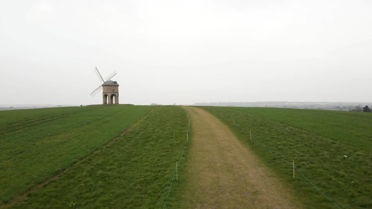 Low aerial swiping shot of an old windmill on a hill surrounded by green fields.