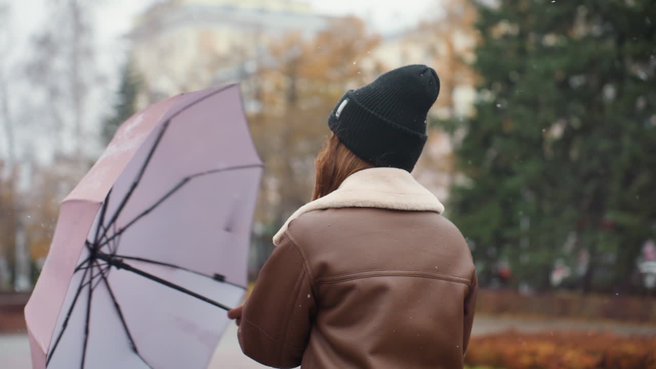 Student holding umbrella, wearing knit cap, brown shearling jacket, happily spinning umbrella in light snowfall, surrounded by trees, enjoying autumn day, joyful and carefree moment outdoors