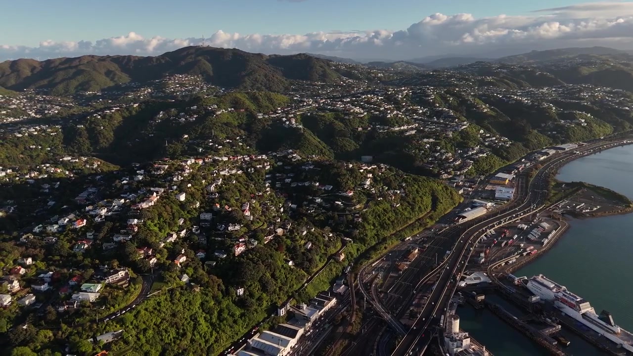 Case a mezza costa scendono a cascata verso una strada portuale curva a Wellington, Nuova Zelanda, incorniciate da fitte colline verdi, un'autostrada costiera e un porto trafficato in lontananza sotto la luce dorata del tramonto. Ampia ripresa aerea con drone.
