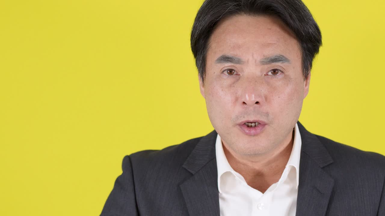 明るい黄色の背景に対して、フォーマルなスーツを着た男性が、唇に指を当てて静止を促す、安定したカメラアングル。