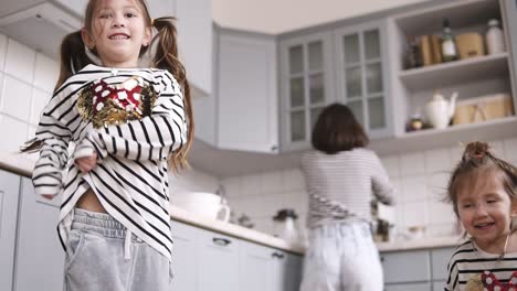 Two-daughters-are-dancing-on-the-kitchen,-mother-are-cooking-behind-them