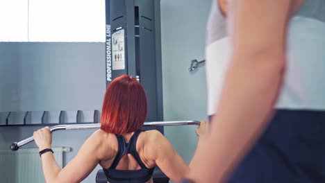 persistent woman doing her workout on a simulator. athletic female trained her muscles pulling down a bar on herself in gym. professional trainer looks at sportwoman's training indoors.