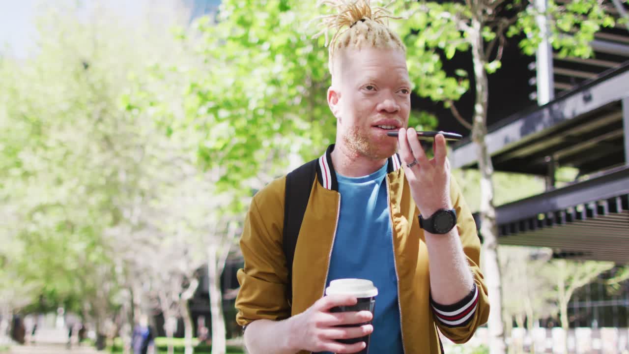 feliz albino hombre afroamericano con rastas en el parque bebiendo café hablando en el teléfono inteligente