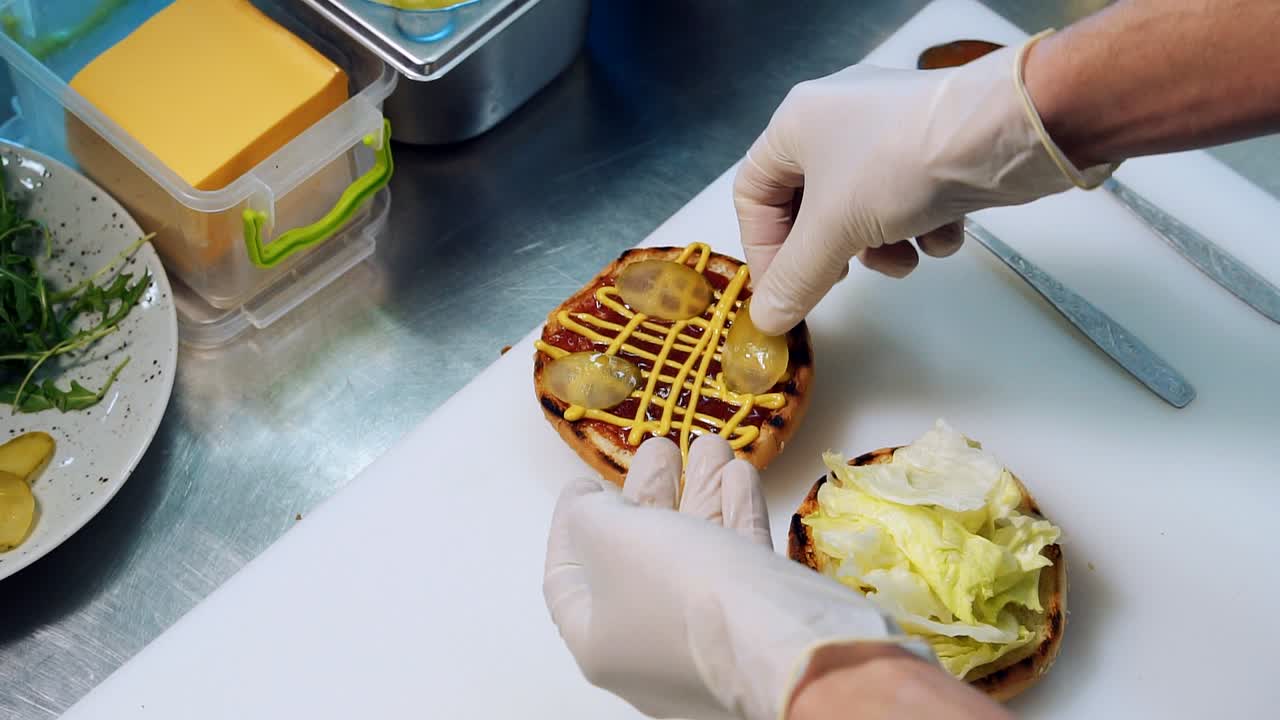 Preparing juicy burger on white board. Chef puts slices of pickled cucumbers on burger bun. Cooking delicious hamburger. Top view