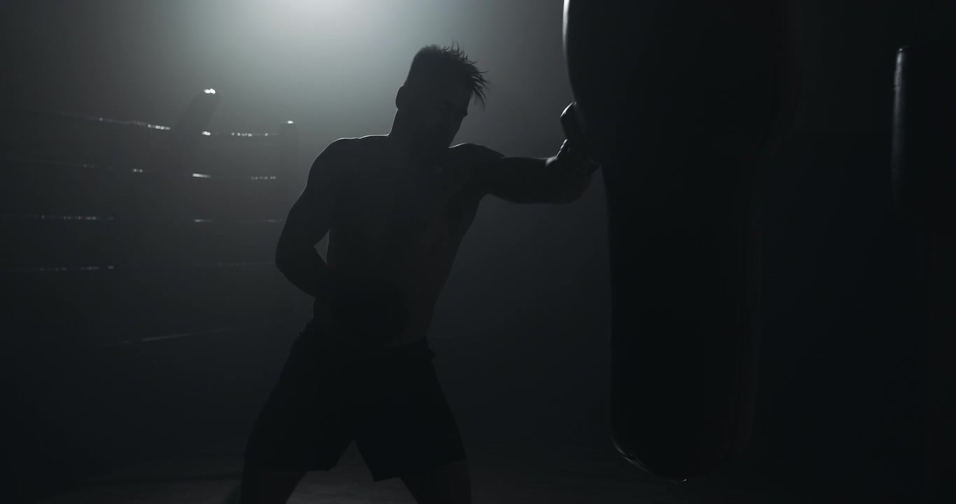Silhouette di pugile che si allena con il sacco da boxe in una palestra buia.