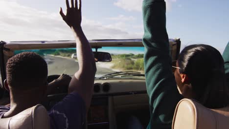 Diverse-couple-driving-on-sunny-day-in-convertible-car-raising-hands-in-the-air-and-smiling