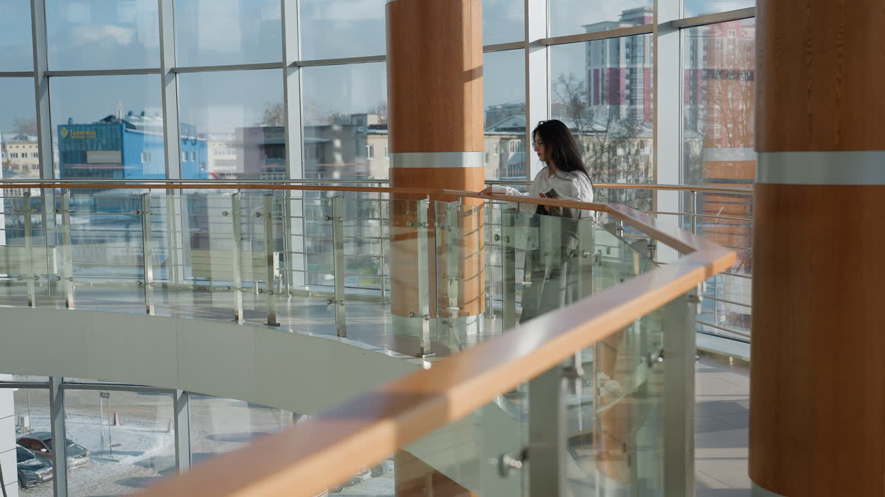 Confident data analyst walking through sunlit mall corridor with suit in hand, pauses by railing and gazes upward thoughtfully, surrounded by glass walls, wooden pillars, and scenic urban skyline