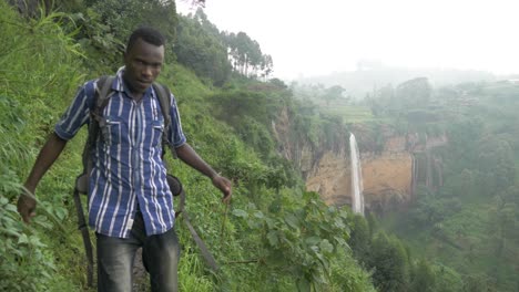 african man walking on a small muddy trail in a tropical jungle with a large waterfall falling off a cliff in the background