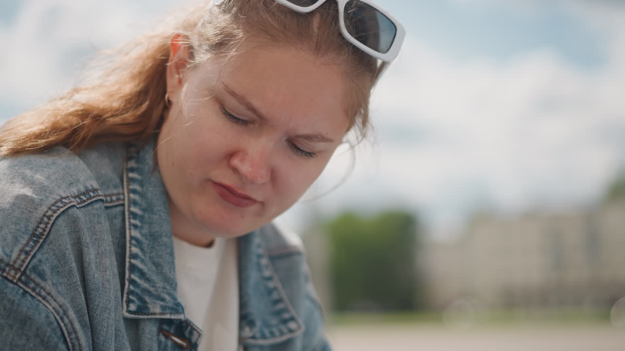 Giovane donna bianca che studia all'aperto, con giacca di jeans e occhiali da sole, seduta su una panchina del parco, concentrata su un quaderno e che prende appunti. Un primo piano spontaneo rivela lentiggini e una coda di cavallo, con luce solare soffusa e sfondo sfocato.
