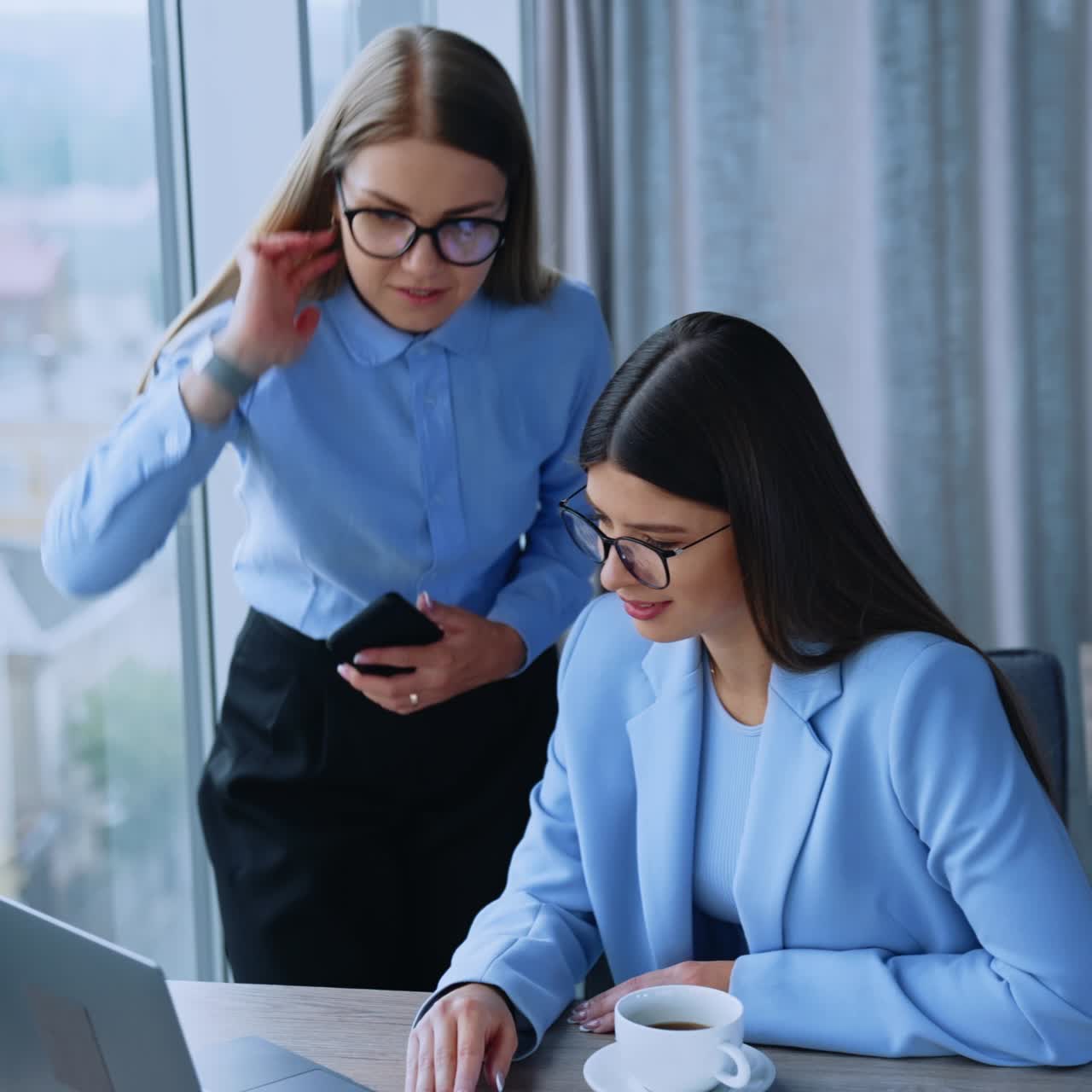 Business communication in the modern office. Female colleagues looking at laptop communicating and smiling. Blurred city panorama in window at backdrop