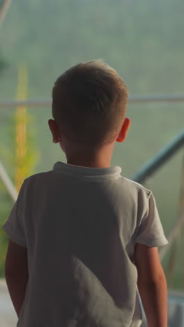 pensive kid examines landscape. preschooler boy approaches big sunlit window in comfortable tent slow motion backside view. calm child admires nature closeup on blurred background