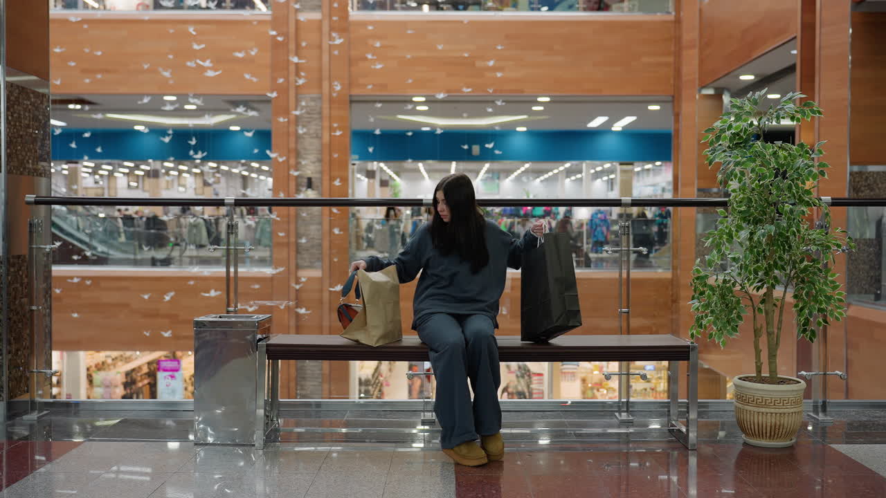 Young girl in dark outfit finishes shopping inside modern mall, drops shopping bag on bench, sits down, crosses leg beside indoor plant and glass railing with view of clothing store behind her