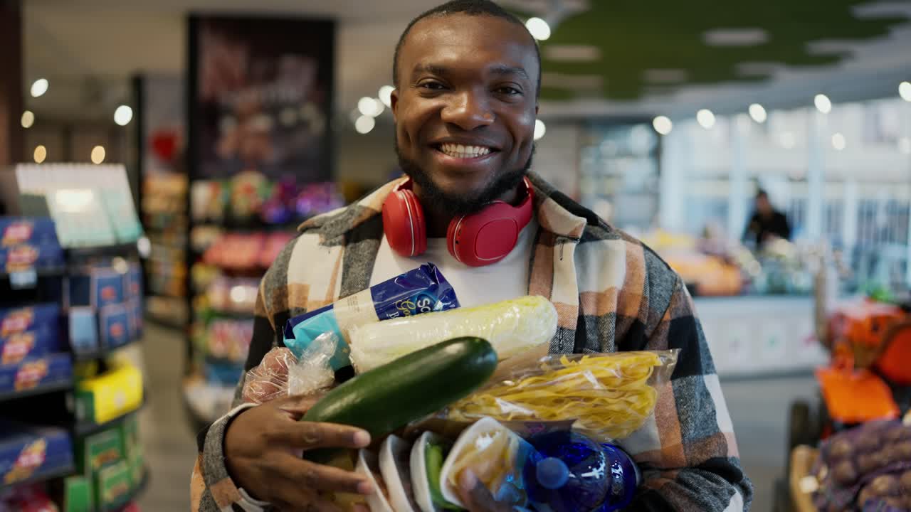 Portrait of a happy man with Black skin in a checkered shirt and red wireless headphones who holds a large amount of products on his chest while holding it with both hands in a modern supermarket near the display cases