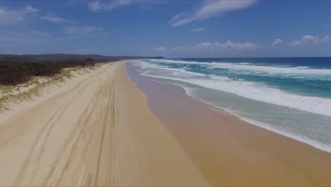 Smooth-and-slow-descending-aerial-drone-shot-looking-North-over-the-dunes,-tire-tracks-and-bushland,-down-a-deserted-Main-Beach,-on-North-Stradbroke-Island-in-Sunny-Queensland,-Australia