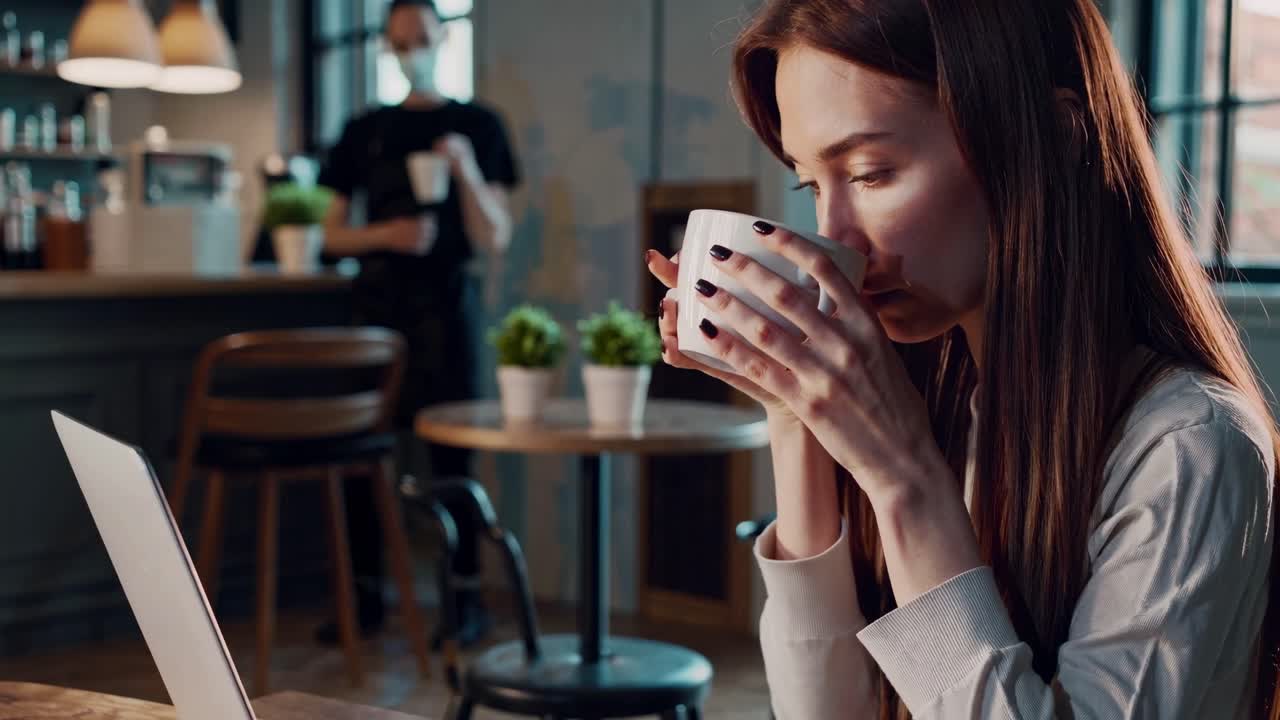 Uma mulher em um café, tomando café e trabalhando em um laptop. O ângulo lateral captura um ambiente aconchegante e relaxante.