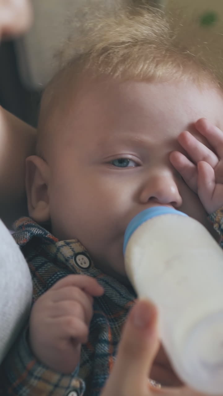 young mommy feeds little tired baby with fresh milk from bottle with dummy in spacious light room close view