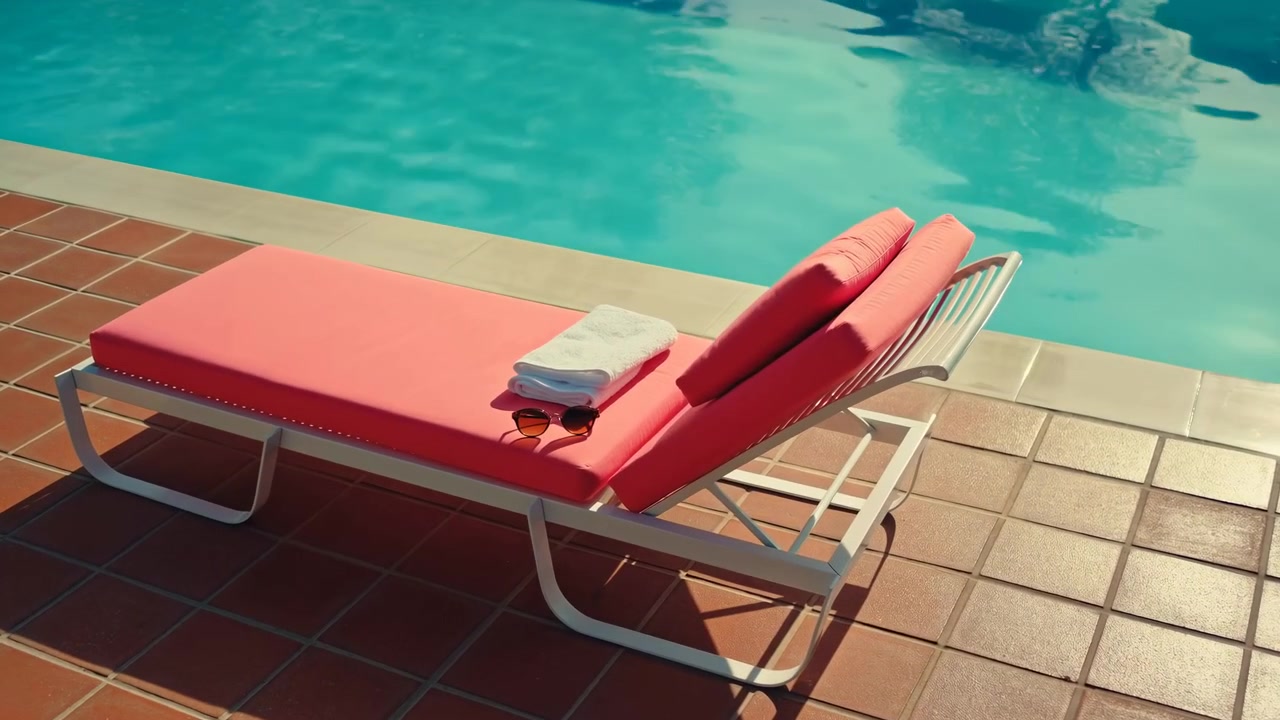 Ein roter gepolsterter Liegestuhl mit gefalteten Handtüchern steht im unteren linken Vordergrund neben einem türkisfarbenen Swimmingpool, mit gefliestem Boden und ruhigem Wasser.