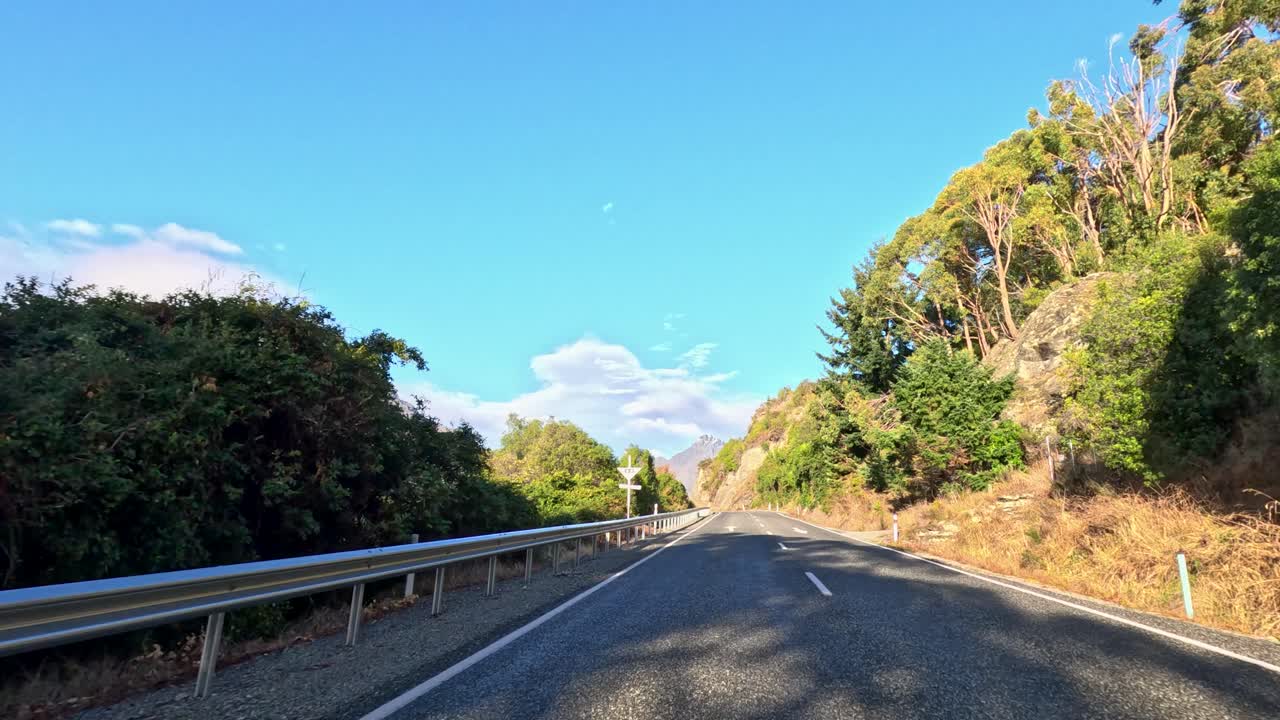 Forward-facing car dashcam captures winding mountain road, lush greenery, bright daylight, and clear sky